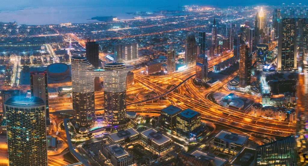 Vue aérienne nocturne de Dubaï, mettant en lumière les gratte-ciel illuminés et l’architecture moderne emblématique de la ville. Paysage urbain vibrant reflétant le dynamisme économique et le développement vertical de la métropole.