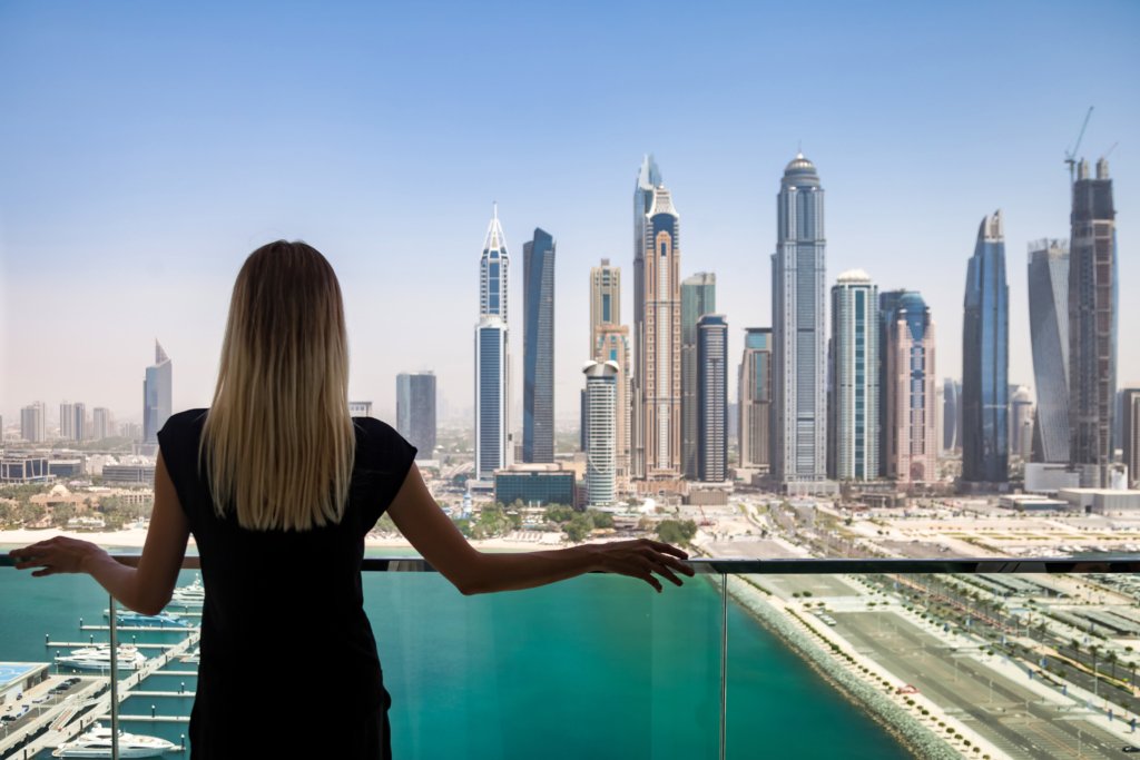Rear view of slim woman in black attire stand on balcony, view of skyscrapers Dubai UAE, looking away. Perfect lady from behind on terrace of tower block. Leisure activity concept.