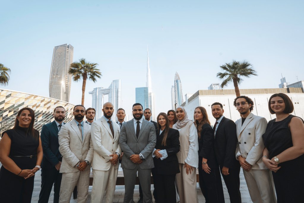 Une quinzaine d'agents immobiliers posent en extérieur, élégamment vêtus, avec le Burj Khalifa et les gratte-ciel de Dubaï en arrière-plan, sous un ciel bleu éclatant.